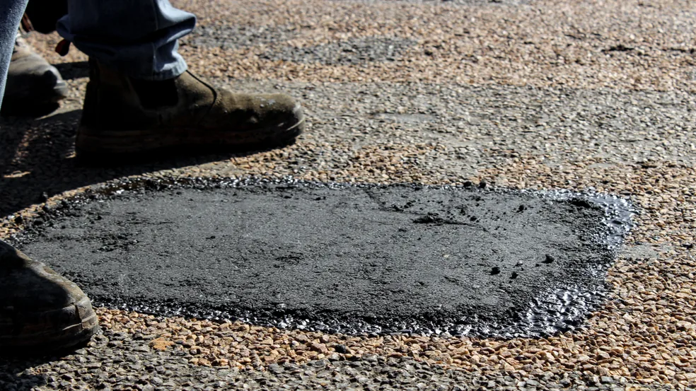 A completed asphalt patch set flush into an older pale aggregate-rich surface, with a worker boots at the edge of the frame