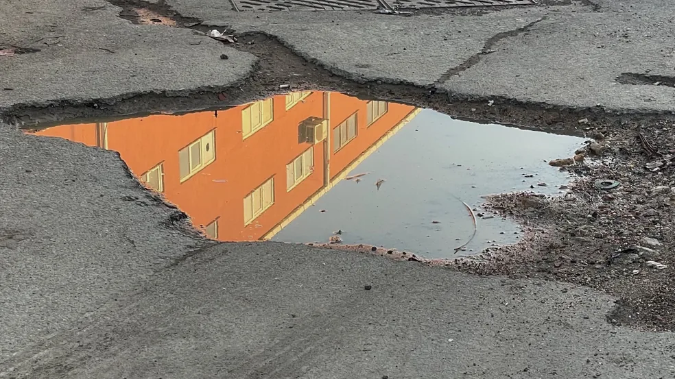 A water-filled pothole in broken asphalt beside a catch basin grate, the surrounding surface split into interconnected cracks