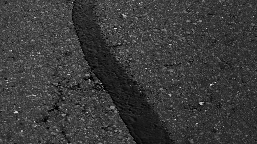 An open crack running across a dark commercial asphalt surface, its edges beginning to break away into loose aggregate